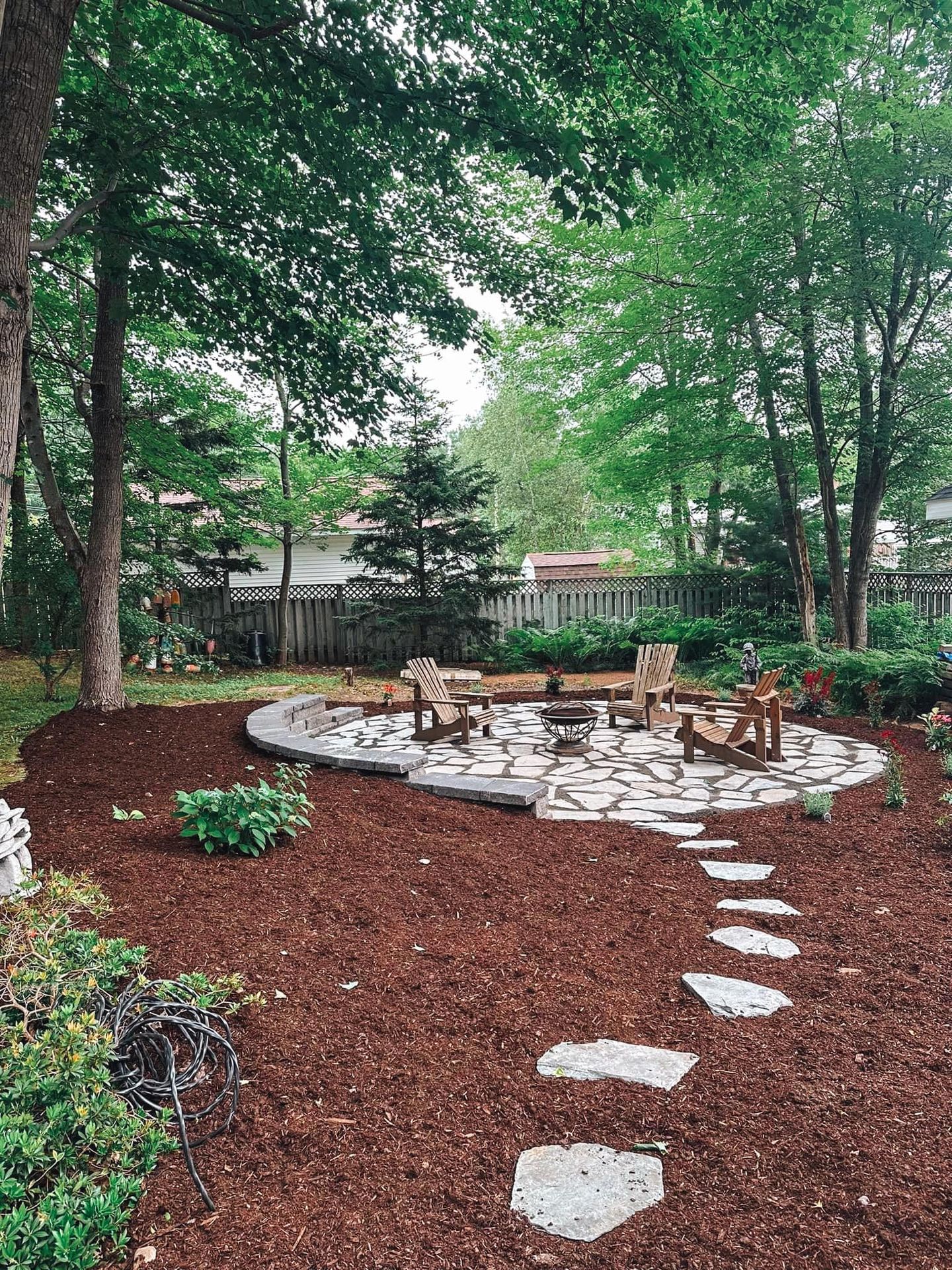 Expertly crafted backyard stone patio with Adirondack chairs and fire pit in Annapolis Valley, Nova Scotia, surrounded by mulch and lush greenery—designed as a serene outdoor retreat by a local landscaping company.
